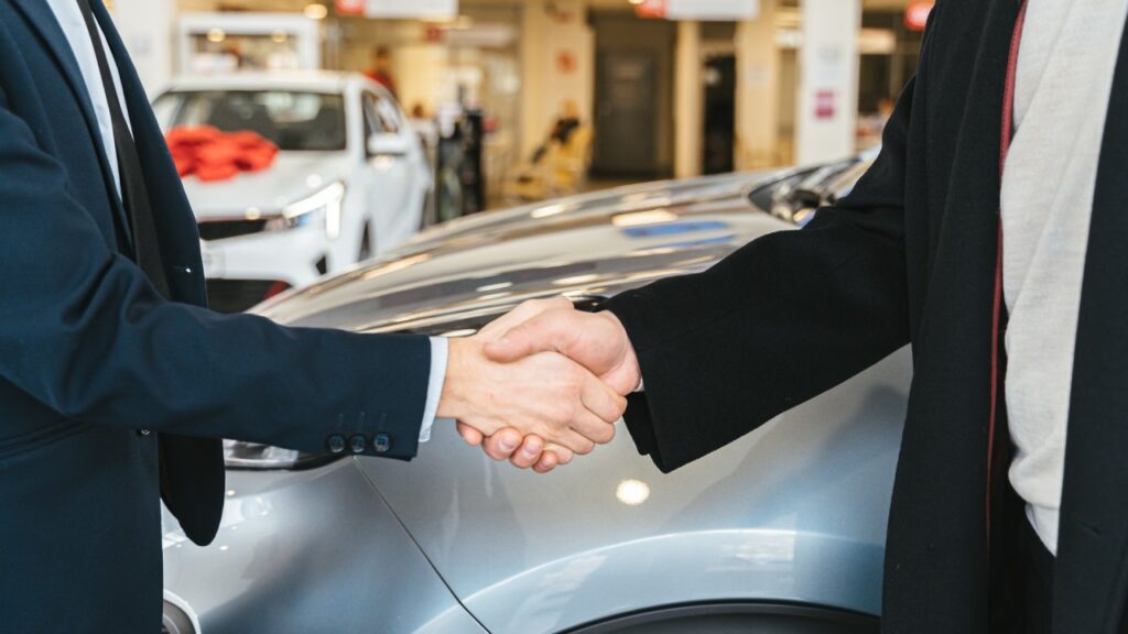 salesperson and customer shaking hands with new car in the background